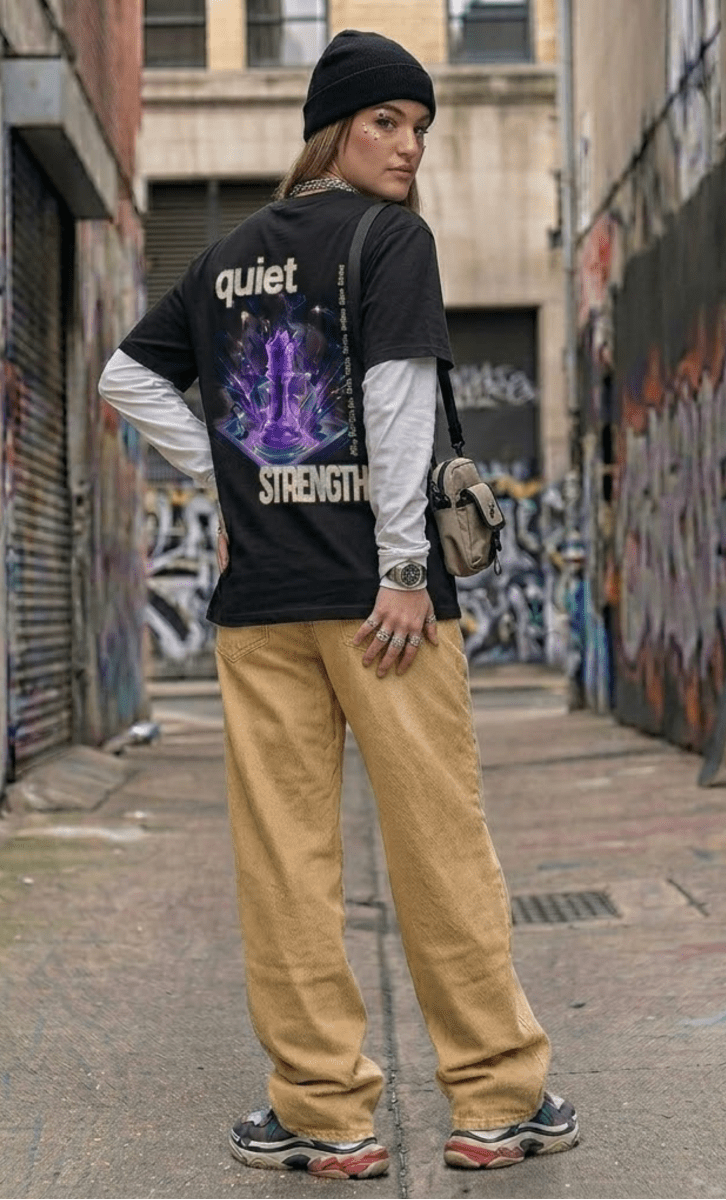 Person wearing a black t-shirt with colorful design and text, standing in an urban alleyway with graffiti.
