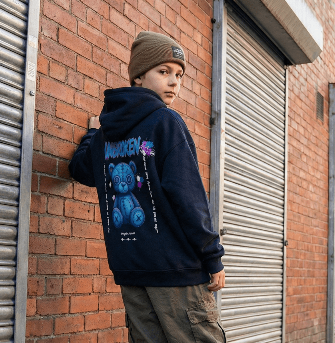 kid wearing a hoodie with a bear graphic leaning against a brick wall.