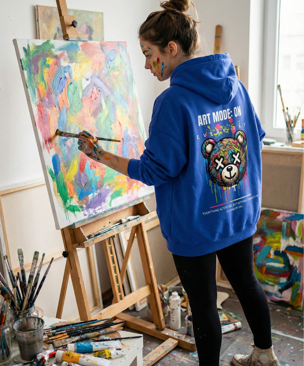 Person painting a colorful abstract artwork in an art studio wearing a blue hoodie with a bear design.
